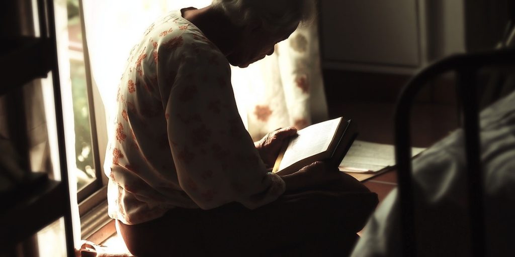Mulher idosa sentada perto da janela lê um livro em silêncio, com o corpo curvado e iluminação quente atravessando a sala.
